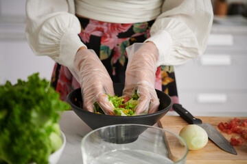 Close-up of young woman in gloves mixing ingredients of vegetable salad in bowl while standing by kitchen table and cooking dinner