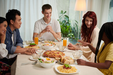 Group of young intercultural people in smart casualwear chatting by served table and enjoying dinner consisting of homemade food