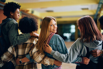 Happy high school student and her friends walk embraced through hallway.