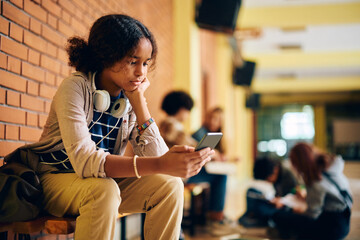Pensive black high school student reads text message on her cell phone in hallway at school.