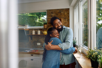Happy black couple in love embraces by window in kitchen.
