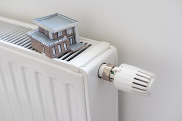 House model wrapped in scarf on radiator indoors, space for text. Winter heating efficiency