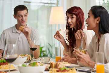 Young woman with snack sitting among her friends by served table during dinner, eating tasty homemade food and discussing something