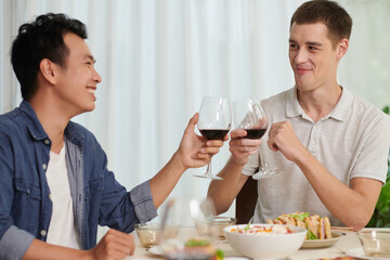 Two young intercultural buddies clinking with glasses of red wine while sitting by table served with homemade food and enjoying dinner