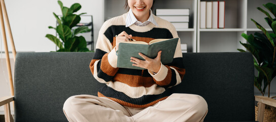 Young Asian woman listening music from headphones and writing note for her work idea in diary book.She in home office or cafe.