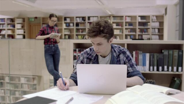 Student boy is working in beautiful new modern library reading room. He is using her laptop computer and typing text data in. Writing a letter, final paper work or just making conspectus for an exam.