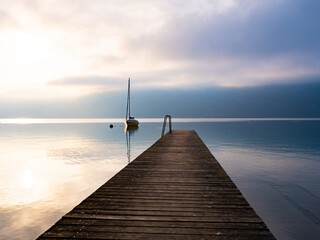 Naklejka na ściany i meble a wooden pier goes into the distance of the lake where the yacht stands against the backdrop of sunrise and mountains in the fog