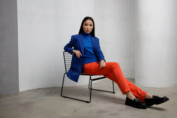 Fototapeta na wymiar High fashion photo of a beautiful elegant young asian woman in pretty blue jacket, blazer, orange pants, trousers. White textured rounded wall