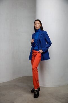 High fashion photo of a beautiful elegant young asian woman in pretty blue jacket, blazer, orange pants, trousers. White textured rounded wall