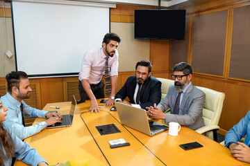 Young indian manager presenting project information to other businesspeople, using laptop and white...