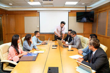 Young indian ceo presenting project information to other businesspeople, using laptop and white...