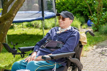 Senior man of 85 years in an electric wheelchair is sitting in a park. Behind him are trees and...