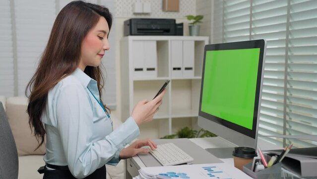 Secretary asian woman in formal dress typing keyboard looking green screen monitor computer desktop and pick mobile phone receive a call in office room. Business person and workplace concept