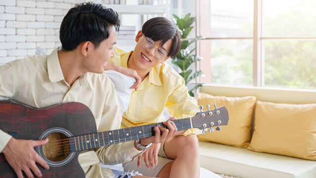 Asian gay couple enjoy spending time together. Man playing a guitar for his partner in the living room.