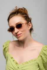 Portrait of young redhead and freckled woman in green blouse and stylish sunglasses standing and posing isolated on grey background, trendy sun protection concept, fashion model