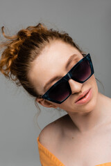 Fototapeta premium Portrait of fashionable young and red haired woman with natural makeup wearing sunglasses while standing isolated on grey background, trendy sun protection concept, fashion model