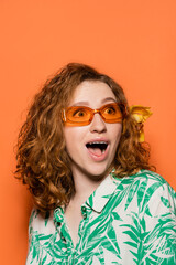 Fototapeta na wymiar Excited young redhead woman with orchid flower in hair and sunglasses looking away while wearing blouse with floral pattern on orange background, summer casual and fashion concept, Youth Culture