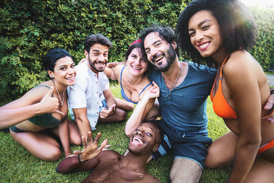 Cheerful and diverse group young friends, capturing a moment of friendship and unity in a selfie They are dressed in swimwear and summer outfits, sitting on a green lawn - Powered by Adobe