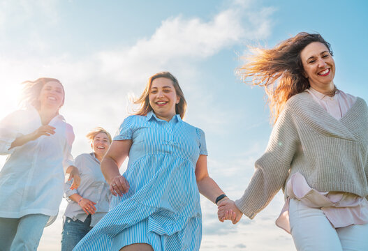Portrait of four cheerful smiling women holding hand in hand running by the meadow. Low angle photo shot on the blue sky with clouds. Woman's friendship, relations, and happiness concept image.