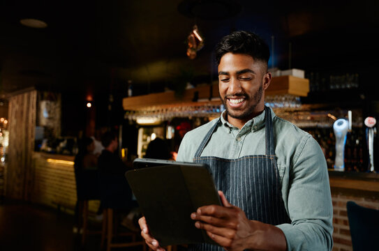 Young biracial man in apron smiling while using digital tablet while working as waiter in restaurant