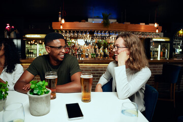 Happy young multiracial group of friends in casual clothing laughing with drinks by table at bar