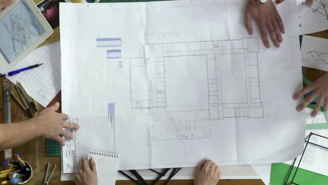 Worktable with building blueprint and architects around it. They analyse information from papers and compare it to the other data on tablet.