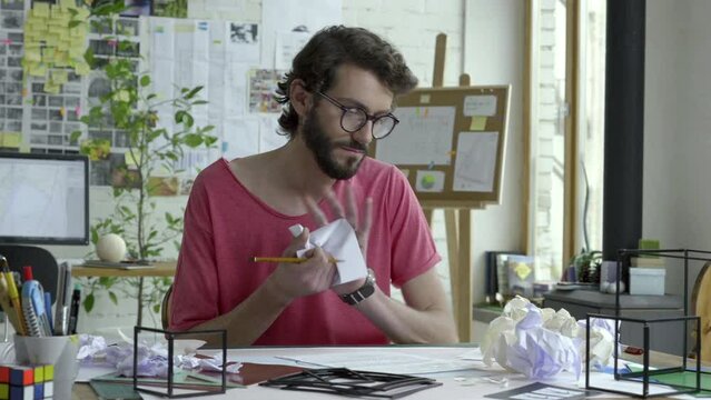 Man is working in his studio or office. He sits near table and write ideas on paper. He is not doing well. He is tired and nervous. He pick new note, write or draw on it, crumple it and throw it away.
