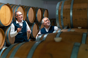Two Professional senior man sommelier tasting and smelling red wine in wine glass at wine cellar with wooden barrel in wine factory. Winery liquor manufacturing industry and winemaker business concept