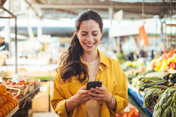 Woman at the farmer's market shopping, sending a text message on her smart phone. Young cheerful...