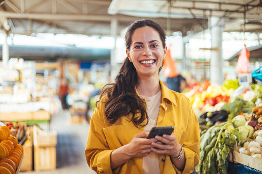 Woman at the farmer's market shopping, sending a text message on her smart phone. Young cheerful woman at the market. Smiling girl decided to cook a delicious and healthy meal