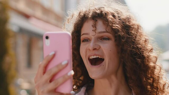 Oh my God Wow. Excited happy joyful winner pretty young woman use smartphone typing browsing celebrating win good message news. Girl with mobile phone walking in urban sunshine city street outdoors