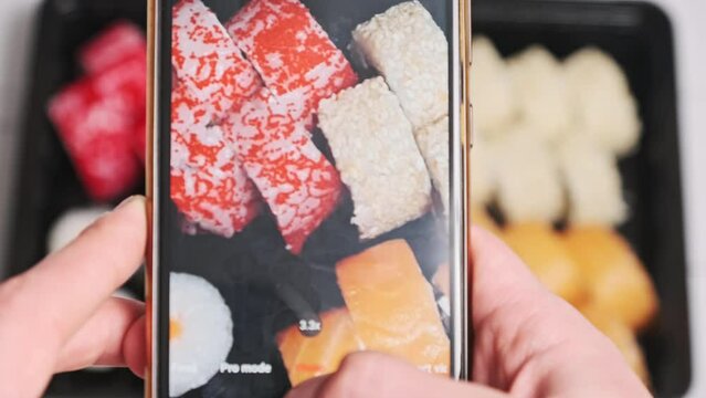 Photographing sushi on a smartphone for social networks. Male hands taking photos of prepared sushi rolls in delivery box on white photo background. Blogger shoots Japanese food on mobile phone camera