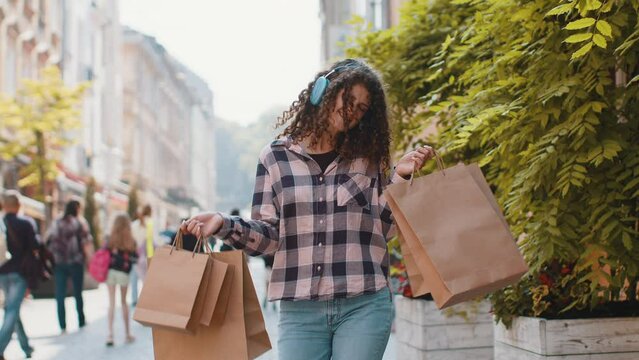 Happy woman in headphones listen music dance after shopping sale with full bags with gifts outdoors. Girl shopaholic consumer tourist traveler walking along the urban city street road. Town lifestyles