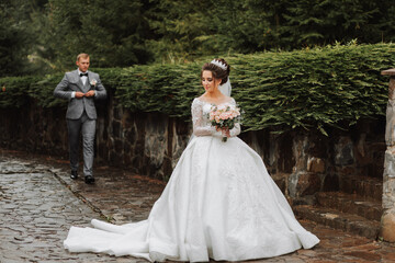 A handsome groom and a charming bride are walking together near the stone fences in the park. Luxurious dress with a long train. groom from behind