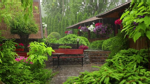 Rain in a picturesque backyard garden with flowers, a bench and a table in summer
