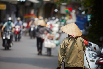 Selective focus on traditional conical hat of person walking against traffic motorbikes on busy street in Old Quarter in Hanoi, Vietnam..