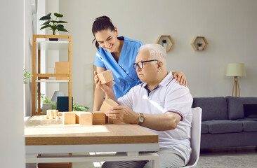 Friendly nurse helping demented old man in retirement home. Senior man with cognitive problems sitting at desk and building wood block house. Dementia care, people with Alzheimer's disease concept