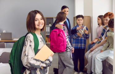 Portrait of smiling girl school girl standing with book and backpack. Happy elementary school student wearing casual clothes posing on background of her classmates in classroom and looking at camera