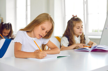 Serious, focused schoolgirl diligently completes task in lesson at elementary school. Girl sits at desk together with her classmate and writes in copybook with pencil. Concept of primary education.