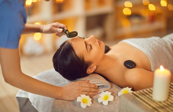 Young Woman Enjoying Face Massage At Spa Salon. Specialist Doing Facial Massage With Green Jade Roller To Beautiful Relaxed Woman Lying On Bed On Towel With Flowers. Beauty, Skin Care Concept