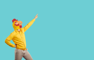 Funny man in chicken rubber head mask isolated on blue studio background make dancer moves. Male in eccentric headwear dance point at empty copy space. Advertising offer concept.