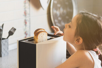Caucasian girl 5 -7 years old cooks breakfast from toast in a toaster in a home kitchen, life style. - obrazy, fototapety, plakaty