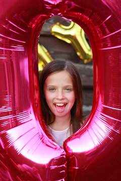 Celebration birthday home party. Funny little girl having fun with big foil pink balloon number 0 and shows painted pink tongue by candy on wooden wall background. Ten years anniversary event.