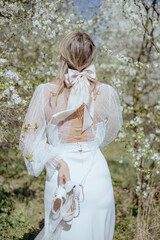 Rear view of young lady in wedding dress standing among cherry trees and hold pair of white high heels shoes with hand.