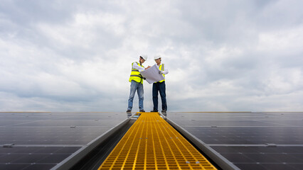 Young Asian engineers discussing, looking at plans for a project to install solar cells on a factory roof, in the foreground are solar panels installed on the factory roof. © VStudio