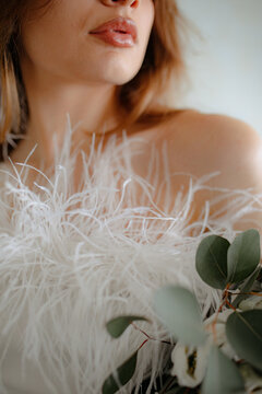 Young brunette woman in shoulderless white clothes adorned with fluffy, little feathers. Lady hold flowers, look away.