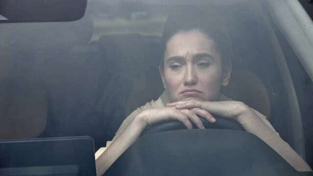 Businesswoman sits in car with sad and upset expression. Black-haired woman scared to drive own car on busy traffic road with mad drivers view through windshield