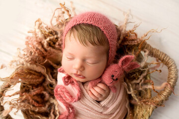 A cute little newborn baby in a white winding and a red cap sleeps sweetly with a red knitted bunny in small hands. Wicker basket. Professional macro photo against a light wooden floor.