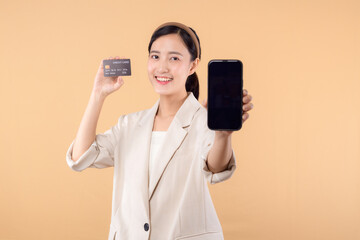 portrait of happy successful confident young asian business woman wearing white jacket holding smartphone and credit card standing over beige background. shopping concept.