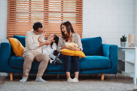 Asian Family Consisting Of Parents, Happy Daughter Watching TV Or Movie Eating Popcorn On Sofa In Living Room At Home. Enjoy Relaxing Happiness.